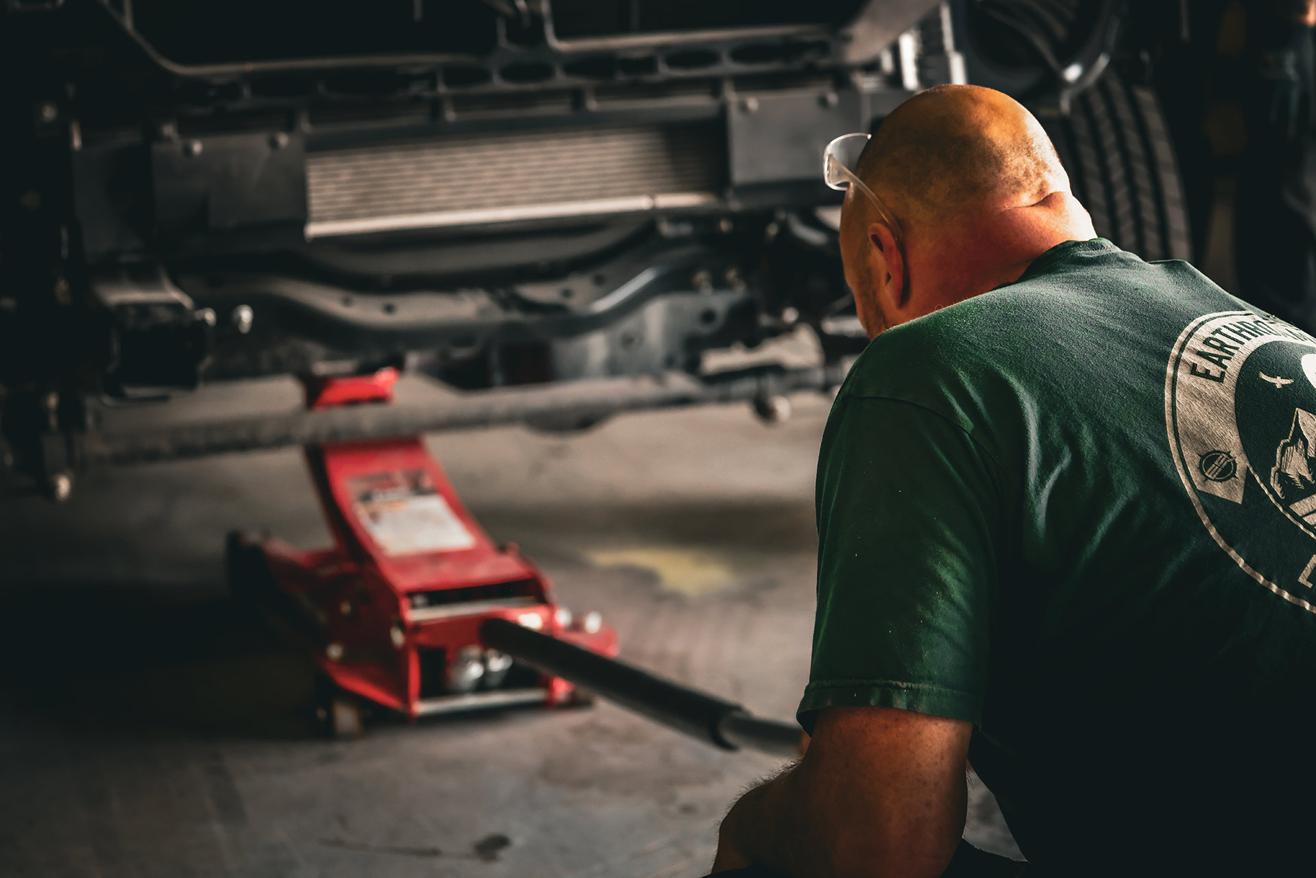 EarthRoamer technician working under a truck with a red hydraulic floor jack in a service bay.